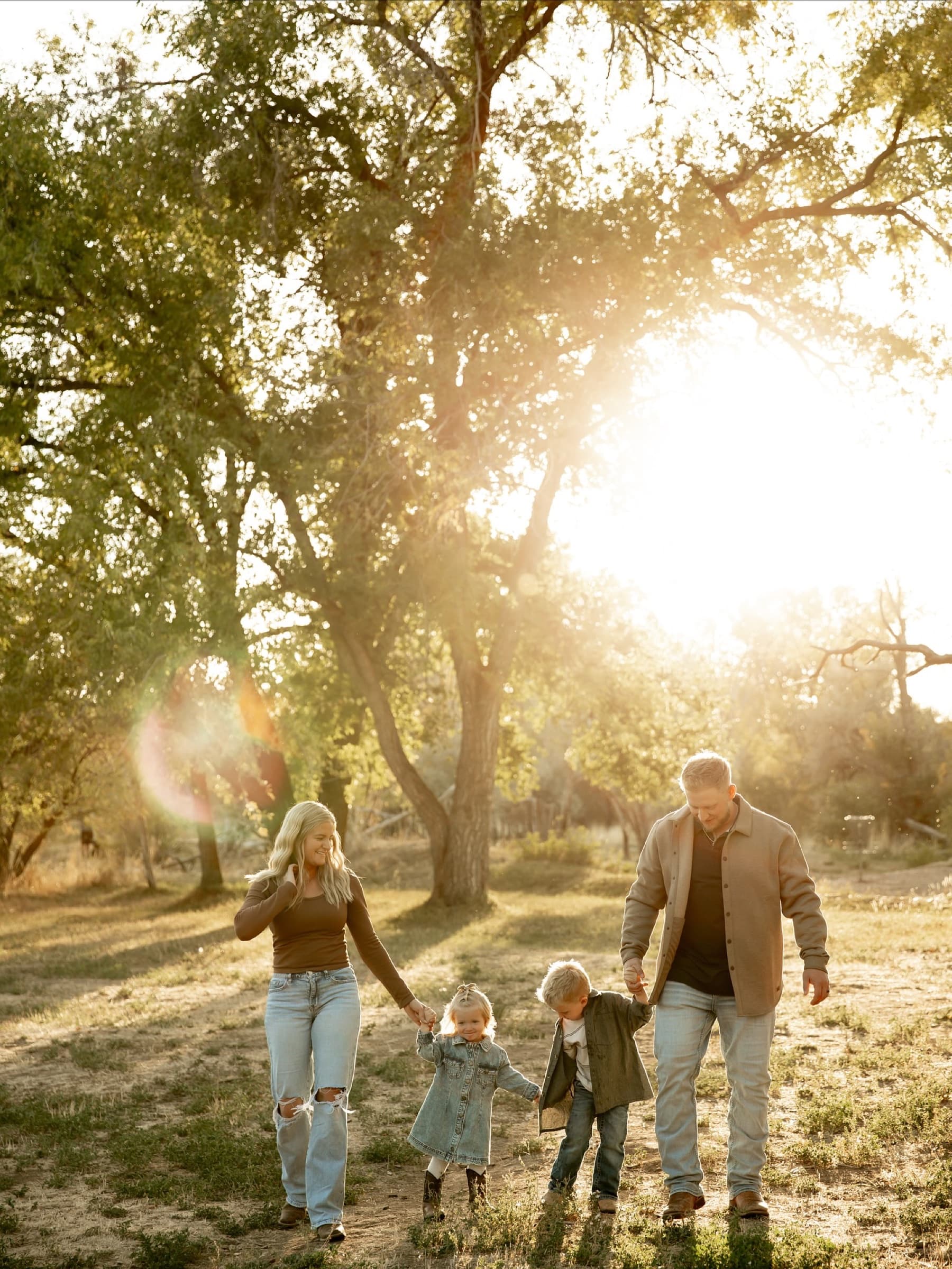 Parents walk hand in hand with their young children beneath tall trees.