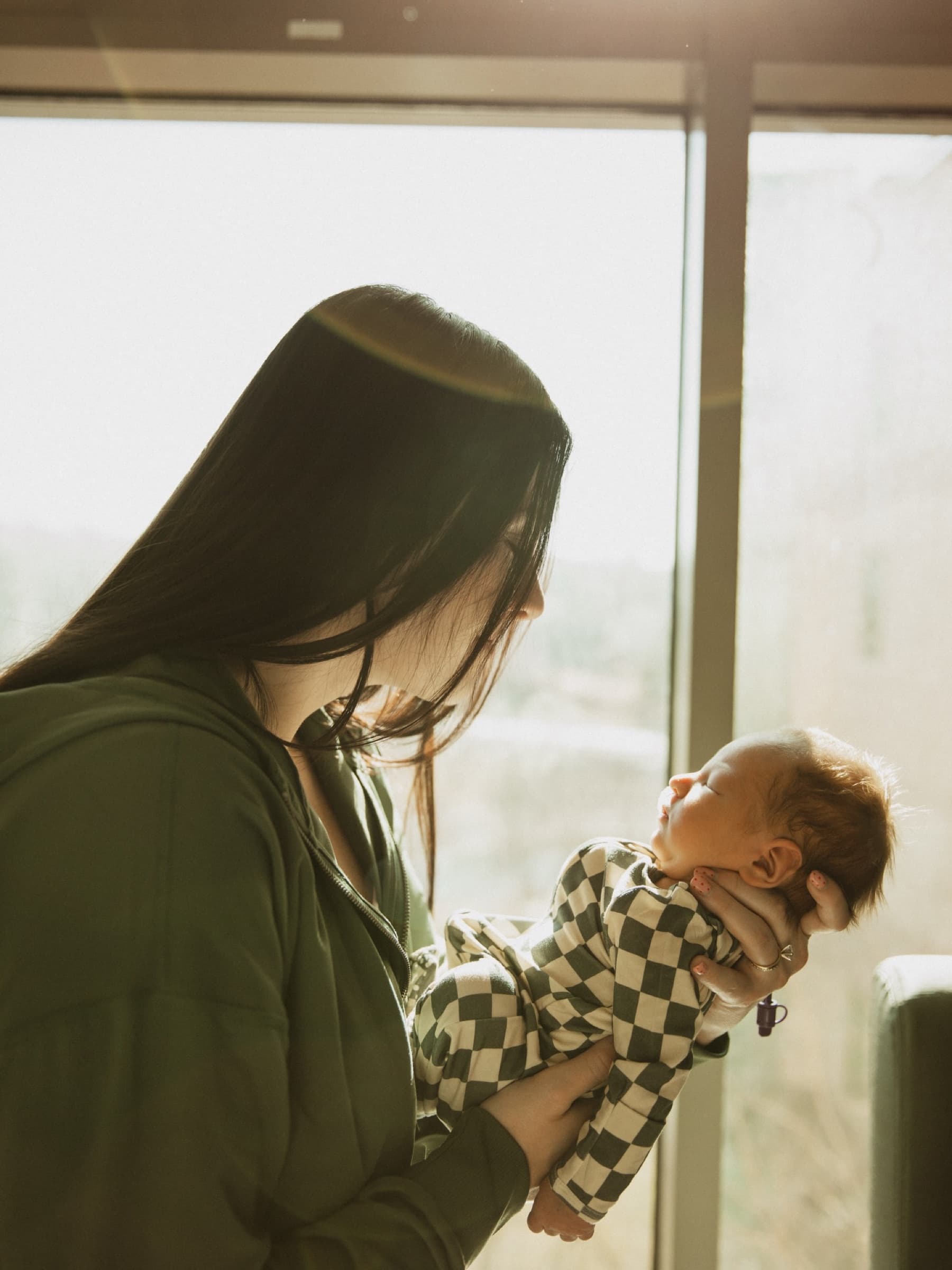 A mother looks at her baby beside a warm window.