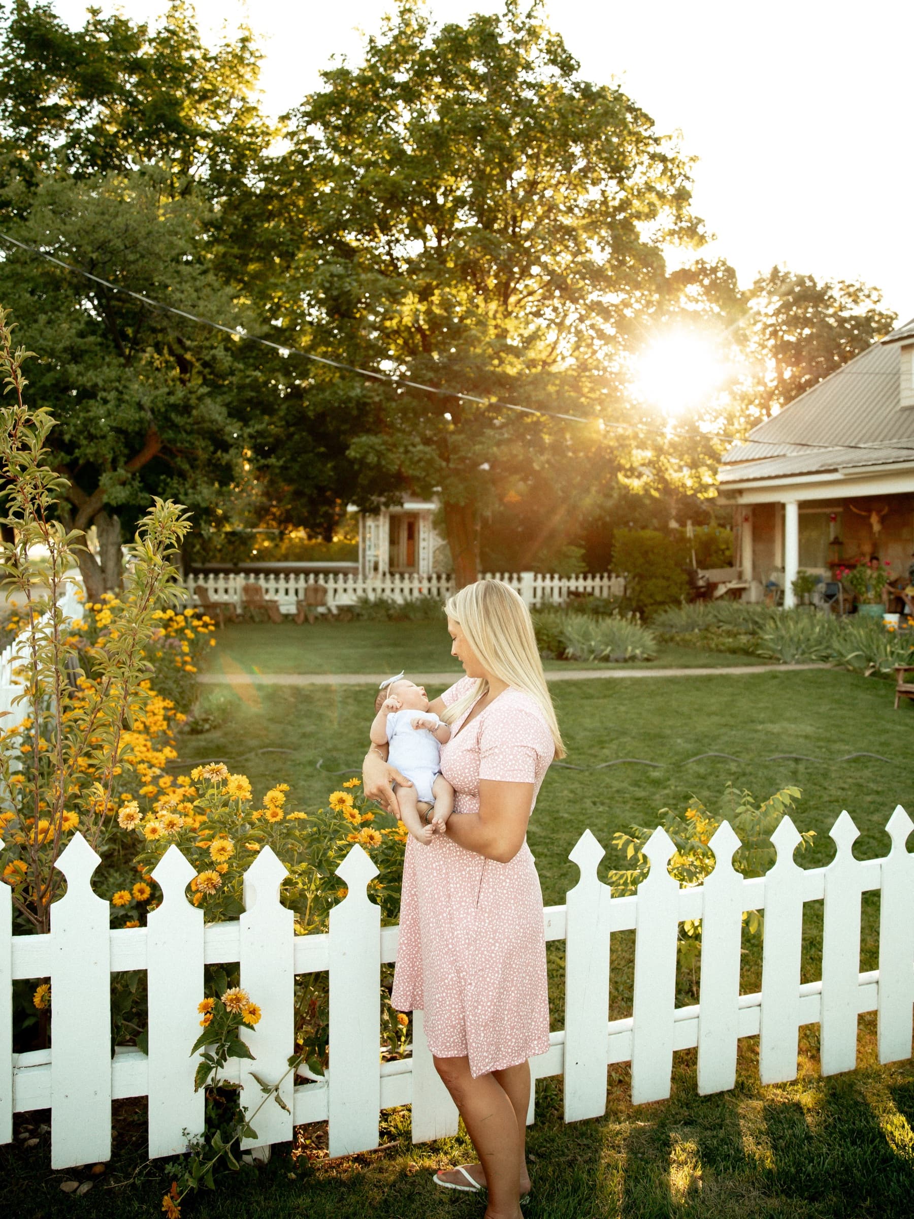 A mother holds her baby beside flowers and a white picket fence.