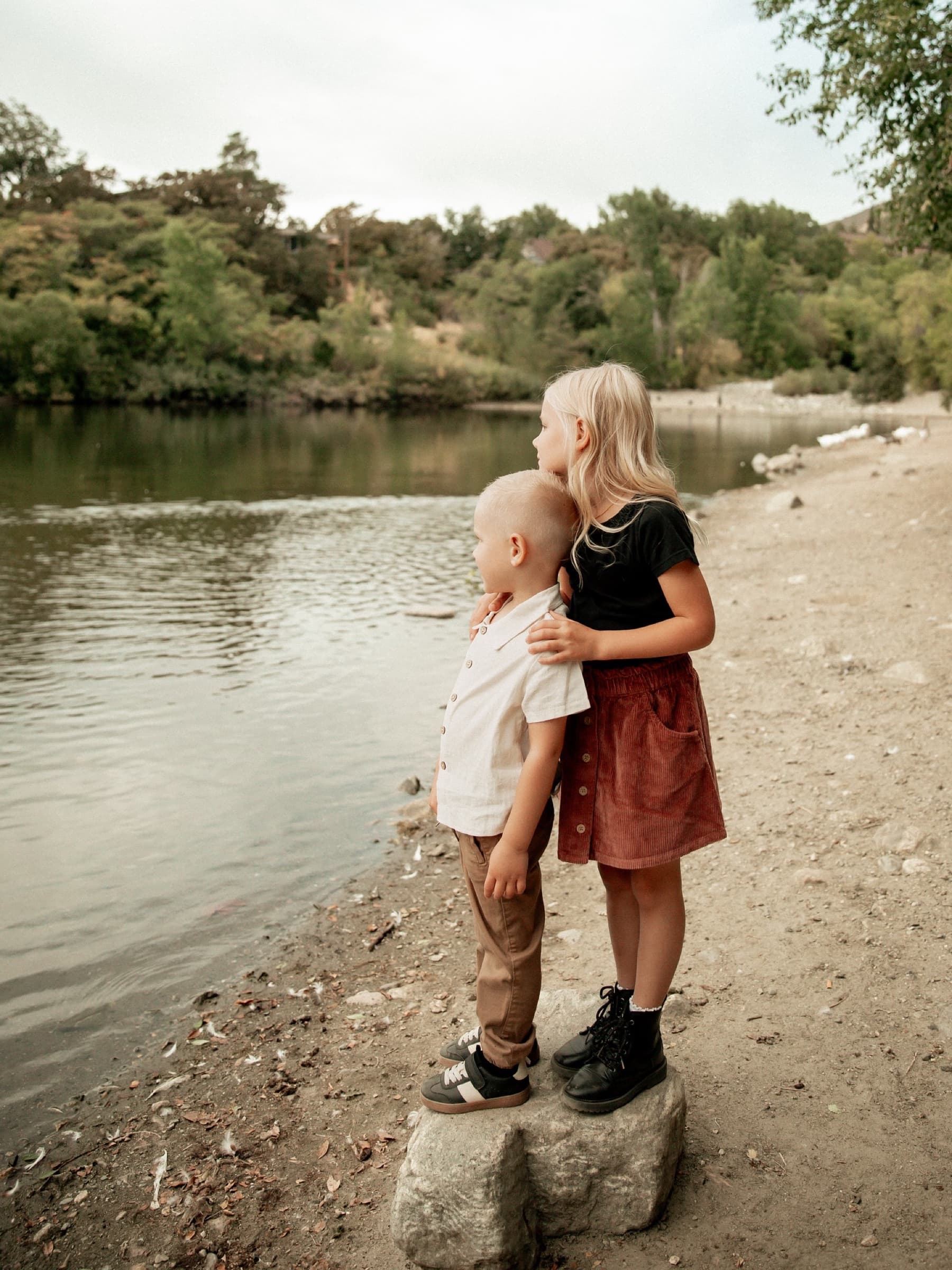 Two siblings stand together at the edge of a quiet river.